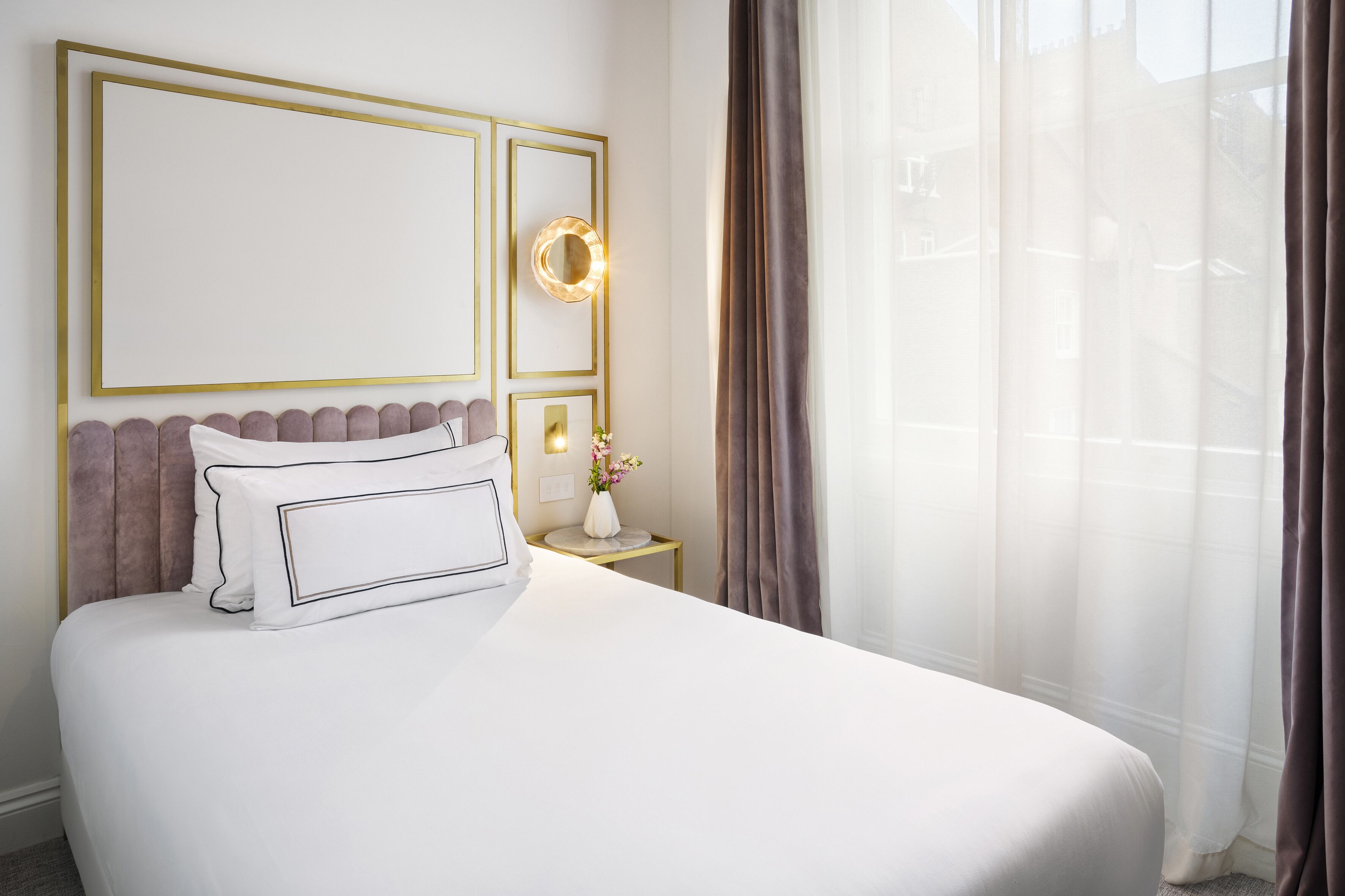 Una cama bien hecha con ropa blanca y cabecera, una mesita de noche con un florero y un ventanal con cortinas translúcidas.