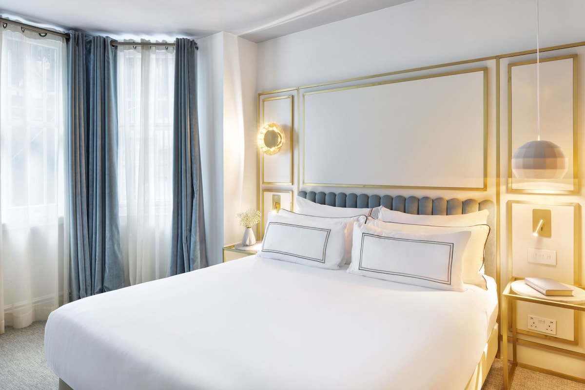 Habitación de hotel con una cama grande, una mesita de noche con lámpara, un espejo con luces y una ventana con cortinas.