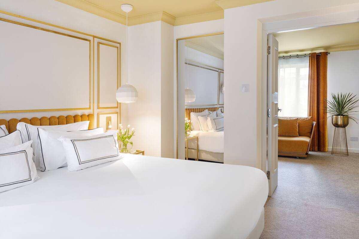 Ein modernes Hotelzimmer mit einem großen Bett, einem Nachttisch mit Lampe und Blick auf ein weiteres Zimmer mit Sofa und Vorhängen.