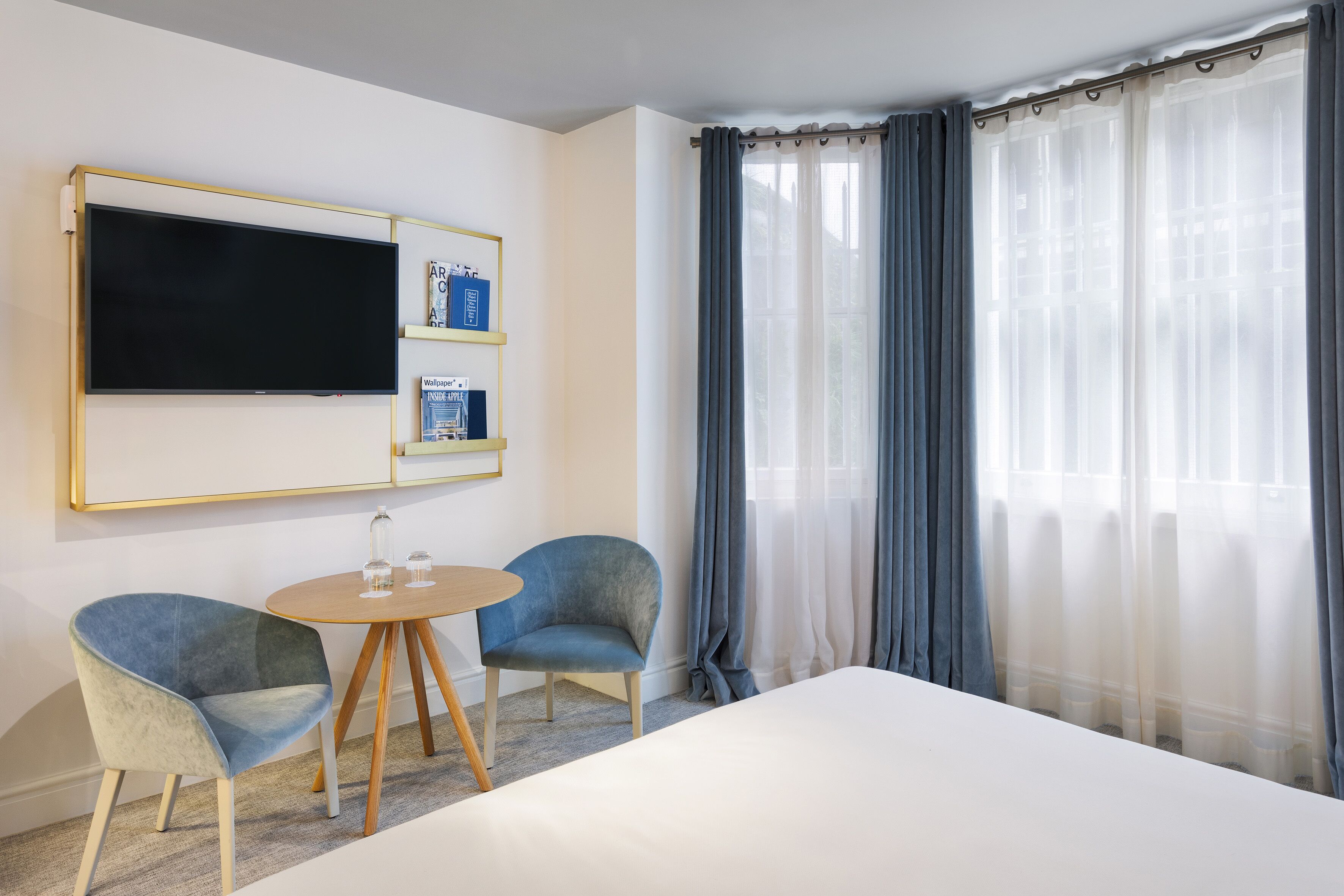 A hotel room with a large bed, two armchairs, a small round table, a flat-screen TV mounted on the wall, and a window with sheer curtains.