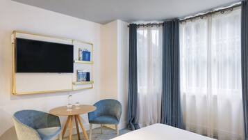A hotel room with a large bed, two armchairs, a small round table, a flat-screen TV mounted on the wall, and a window with sheer curtains.