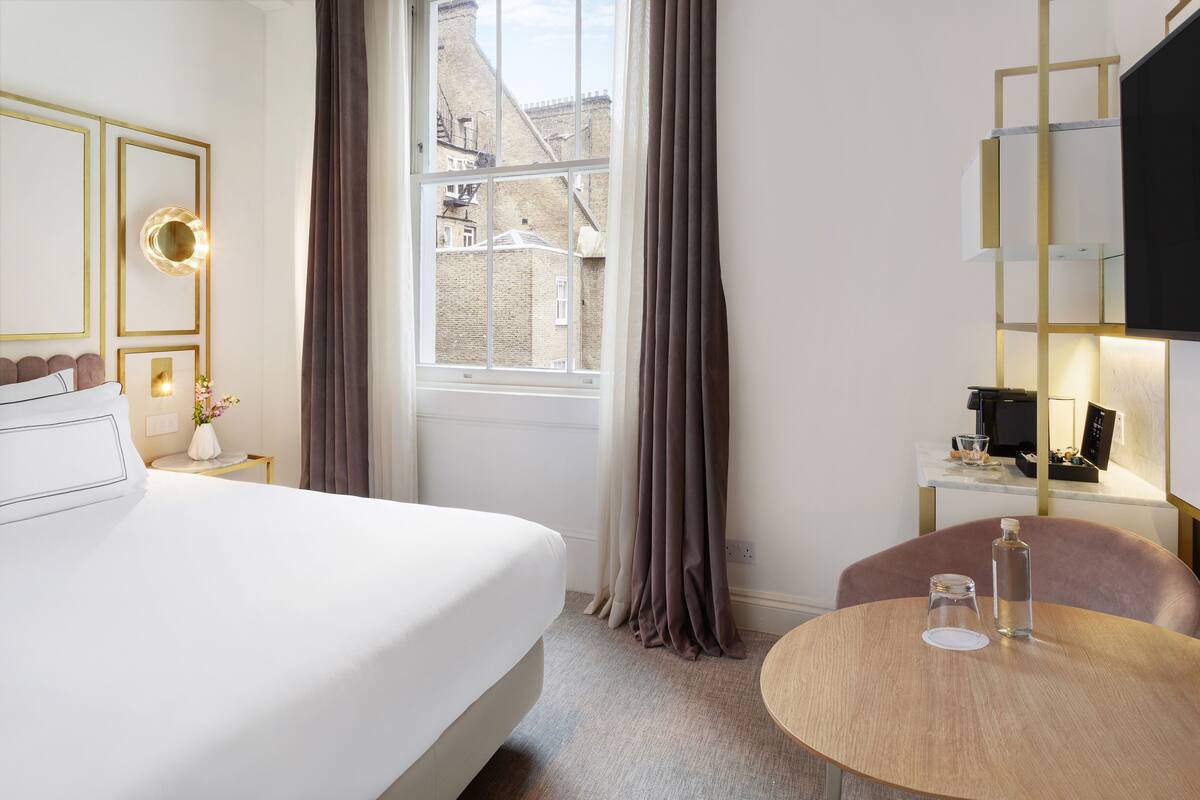 Ein Hotelzimmer mit einem Bett, einem runden Tisch mit Flasche und Glas, einem Schreibtisch mit Computer sowie Blick auf Gebäude durch das Fenster.