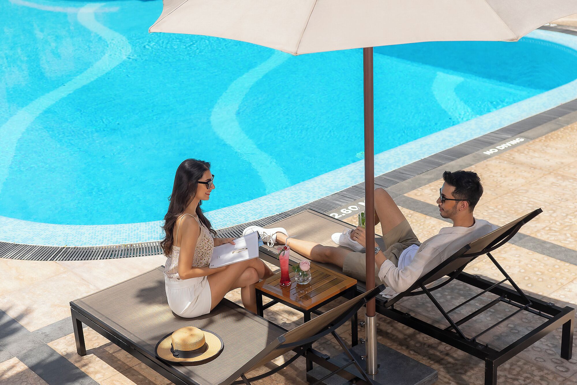 Una pareja descansando junto a una tumbona con una mesa, un sombrero y una bebida.