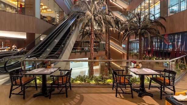 A modern indoor atrium with a central escalator, palm trees, and a dining area with tables and chairs. - The Landmark Bangkok (Bangkok)