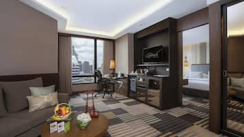 A modern hotel room with a sofa, a dining area, a kitchen, and a large TV.