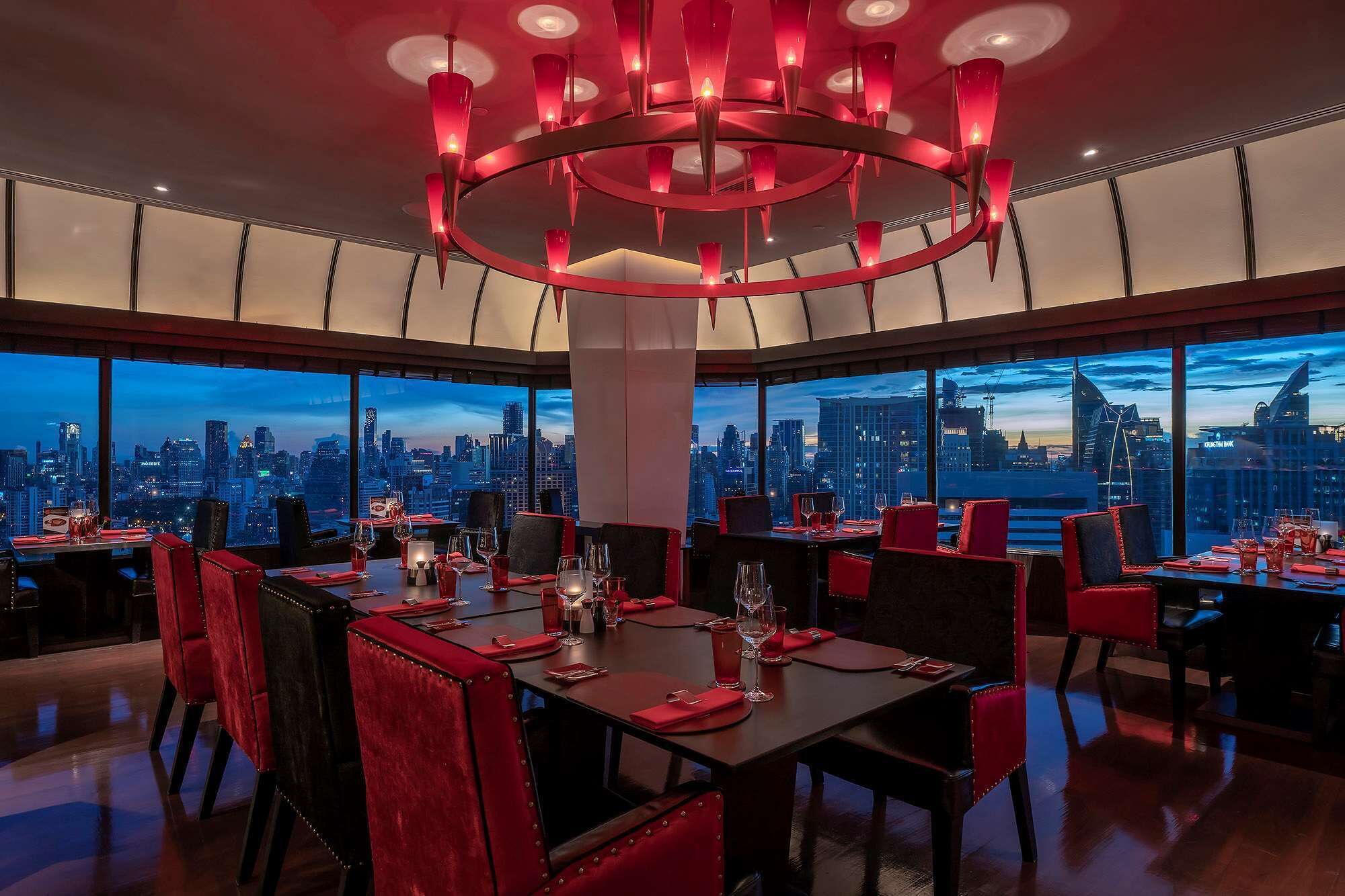 A restaurant with a city skyline view, modern chandelier, and red upholstered chairs.