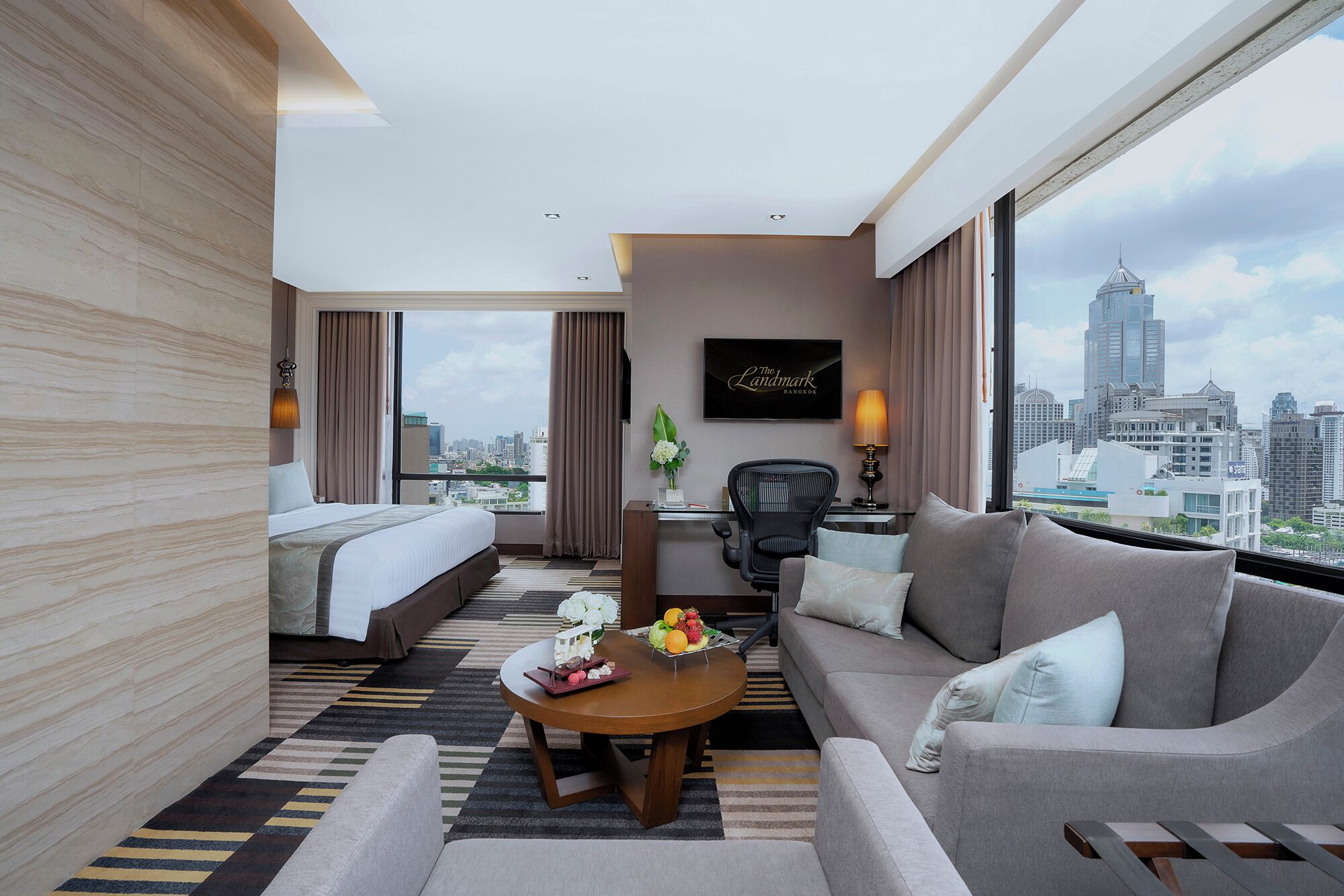 A hotel room with a bed, sofa, and a desk with a lamp. There is a city view through large windows.