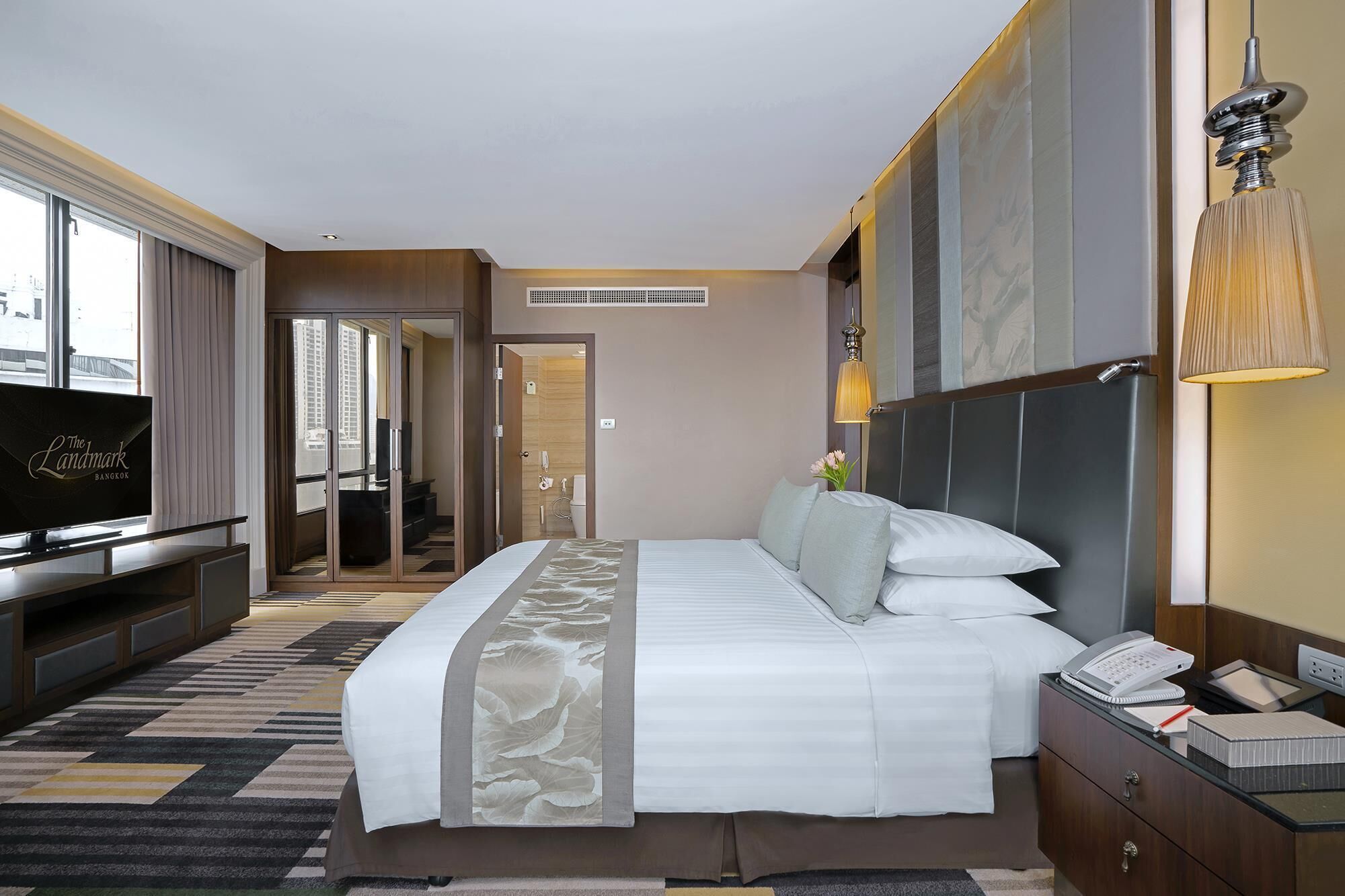 A hotel room with a bed, bedside table, lamp, TV, and a view of the city.