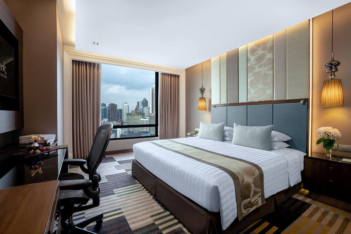 A hotel room with a large bed, a desk, and a chair. There is a view of the city through the window.