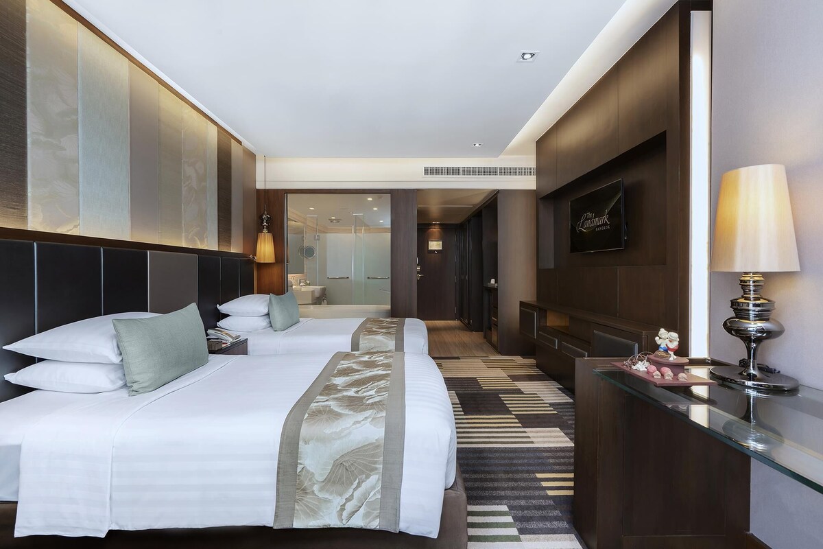 A hotel room with two beds, a glass partition, a bathroom, and a TV.