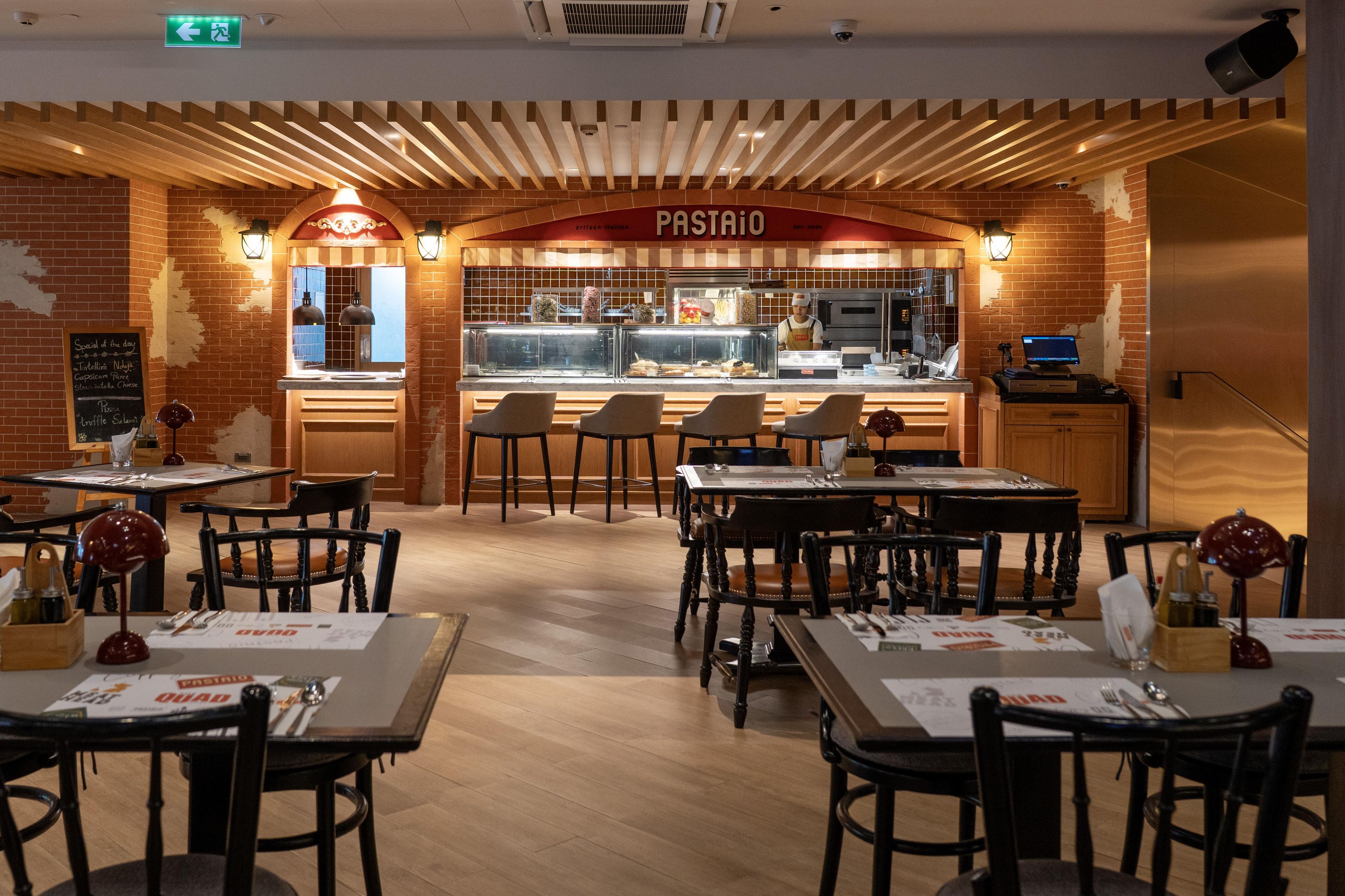 A restaurant interior with brick walls, wooden ceiling beams, and a central counter with a menu display. Tables and chairs are arranged for dining.