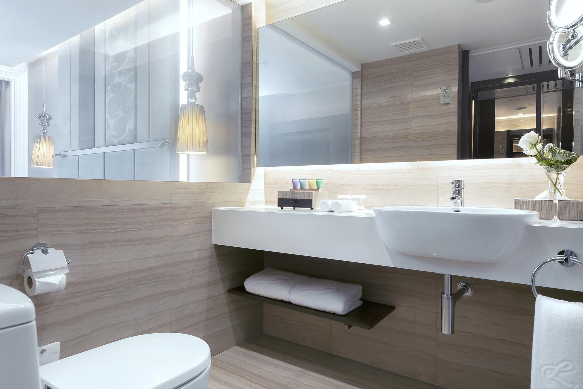 Un baño moderno con lavamanos, espejo y retrete blancos. Hay toallas y artículos de tocador sobre el mostrador.