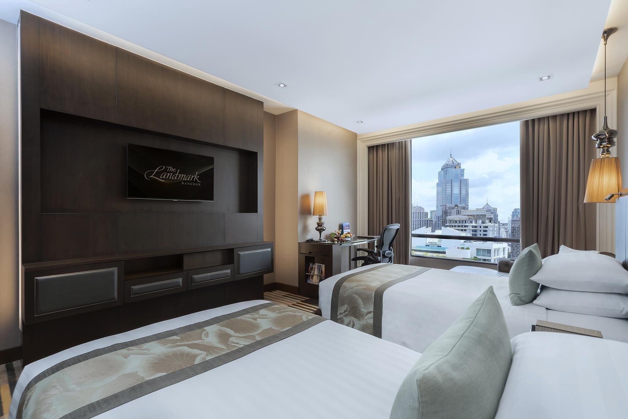 A hotel room with two beds, a TV mounted on the wall, a desk with a lamp, and a view of the city skyline.