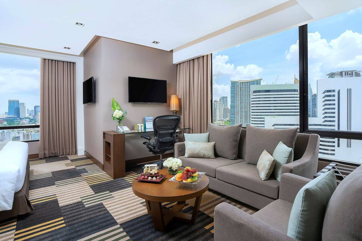 A hotel room with a bed, a sofa, a coffee table with fruit, a desk with a phone, and a TV on the wall.