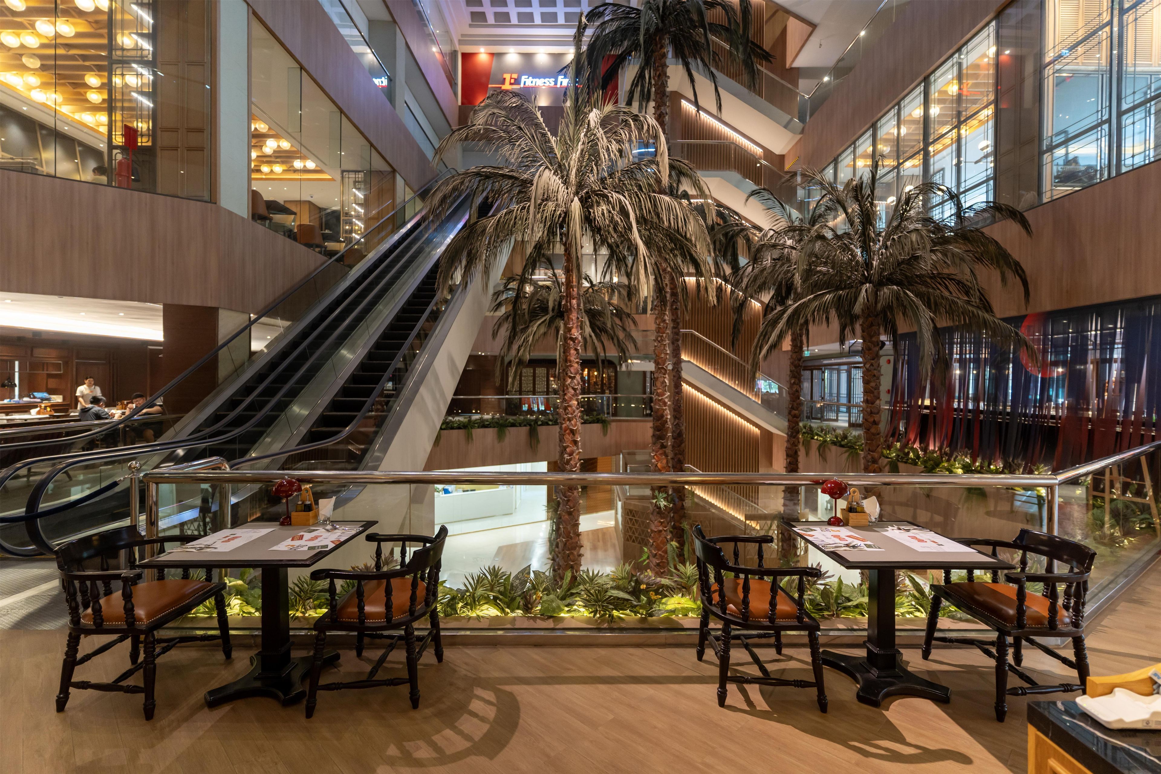 A modern indoor atrium with an escalator, palm trees, and seating areas with tables and chairs.