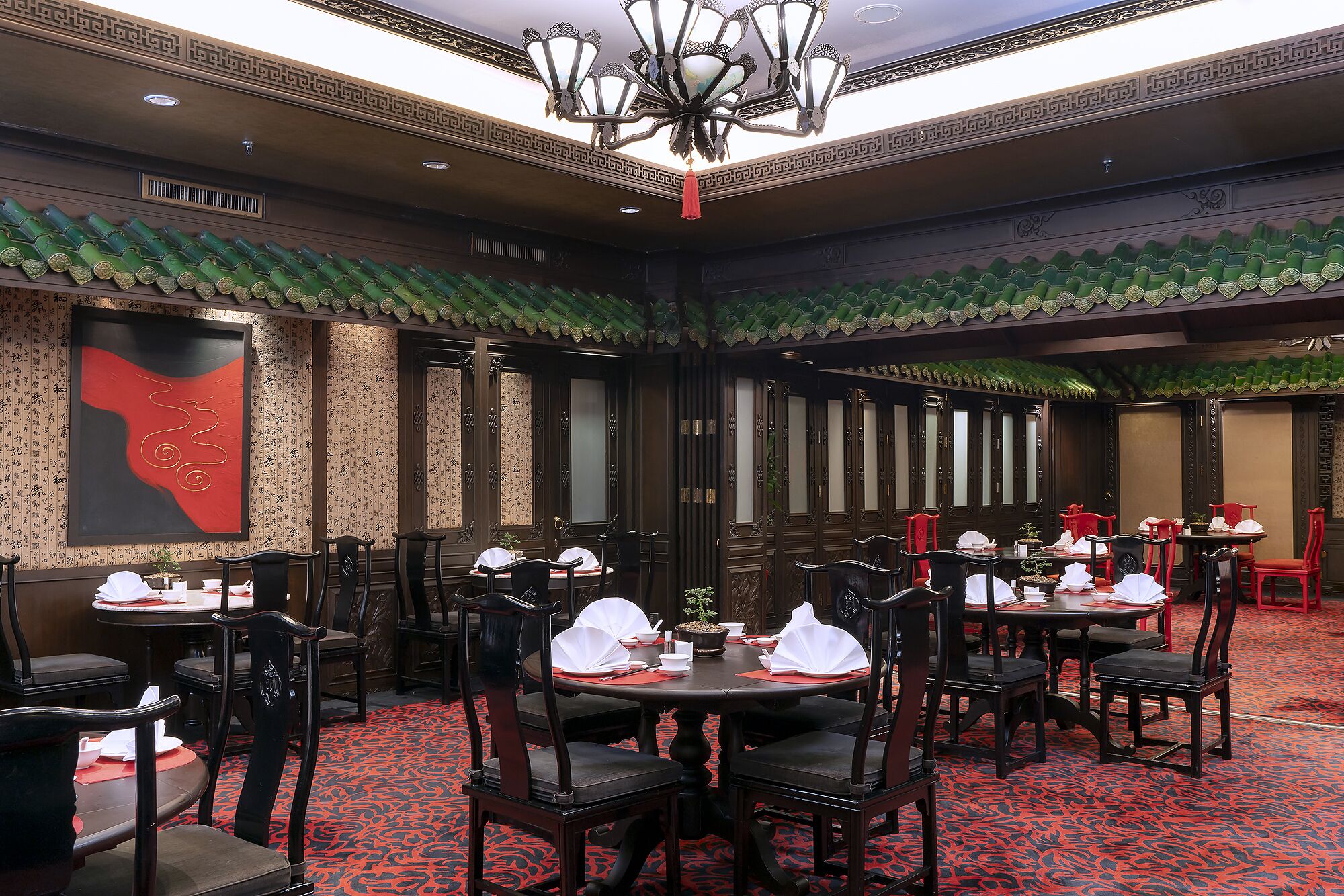 A restaurant with traditional decor, including patterned wallpaper and green-tiled roofs. Tables are set with white tablecloths and red placemats.