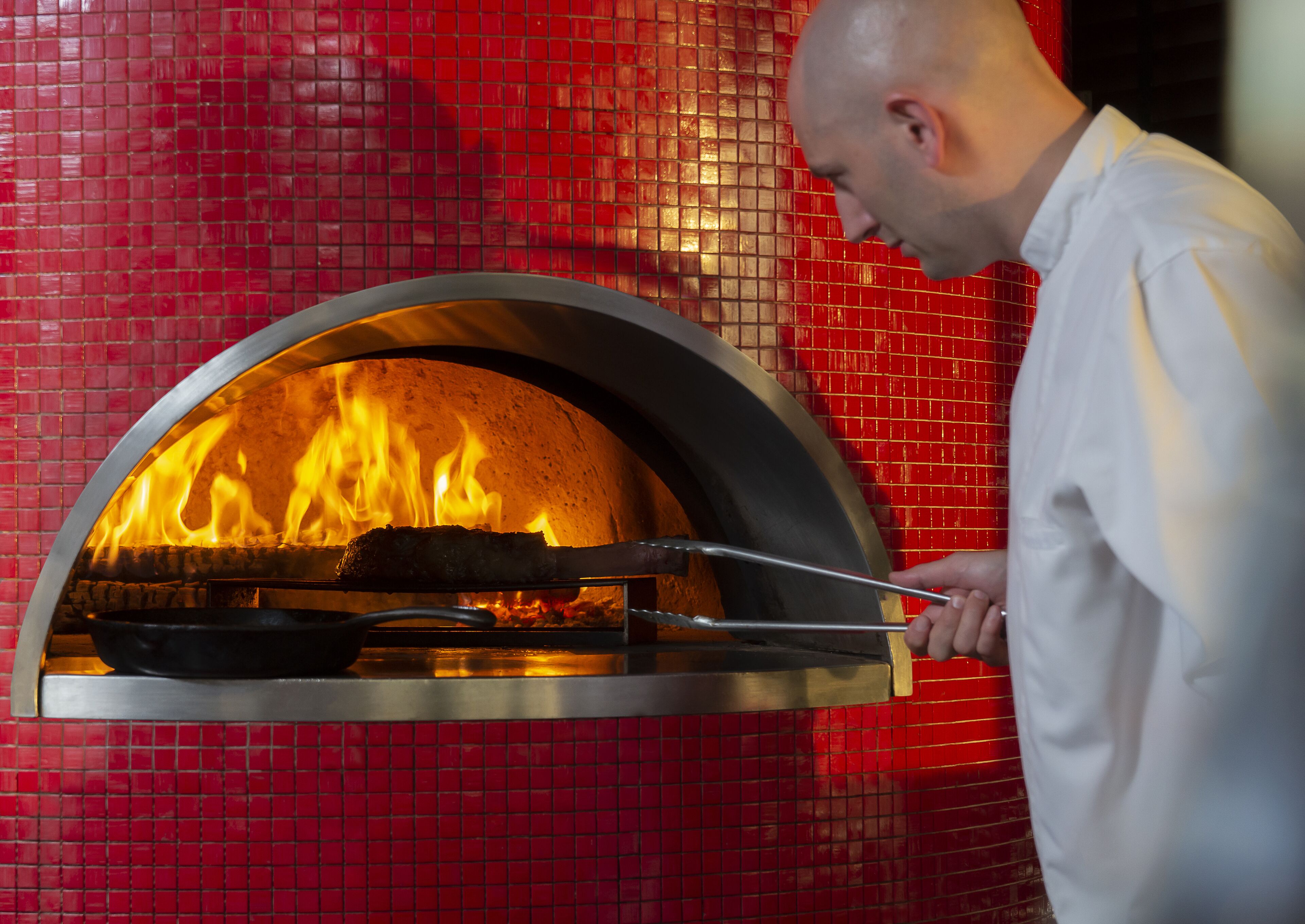 Henkilö, jolla on valkoinen työasu, käyttää pihdejä asettaakseen pizzan näkyvien liekkien keskellä olevaan tiiliuuniin.