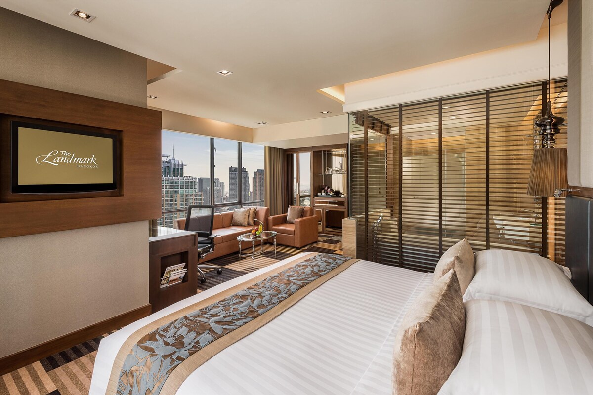 A modern hotel room with a large bed, a sofa, a desk, and a view of the city.