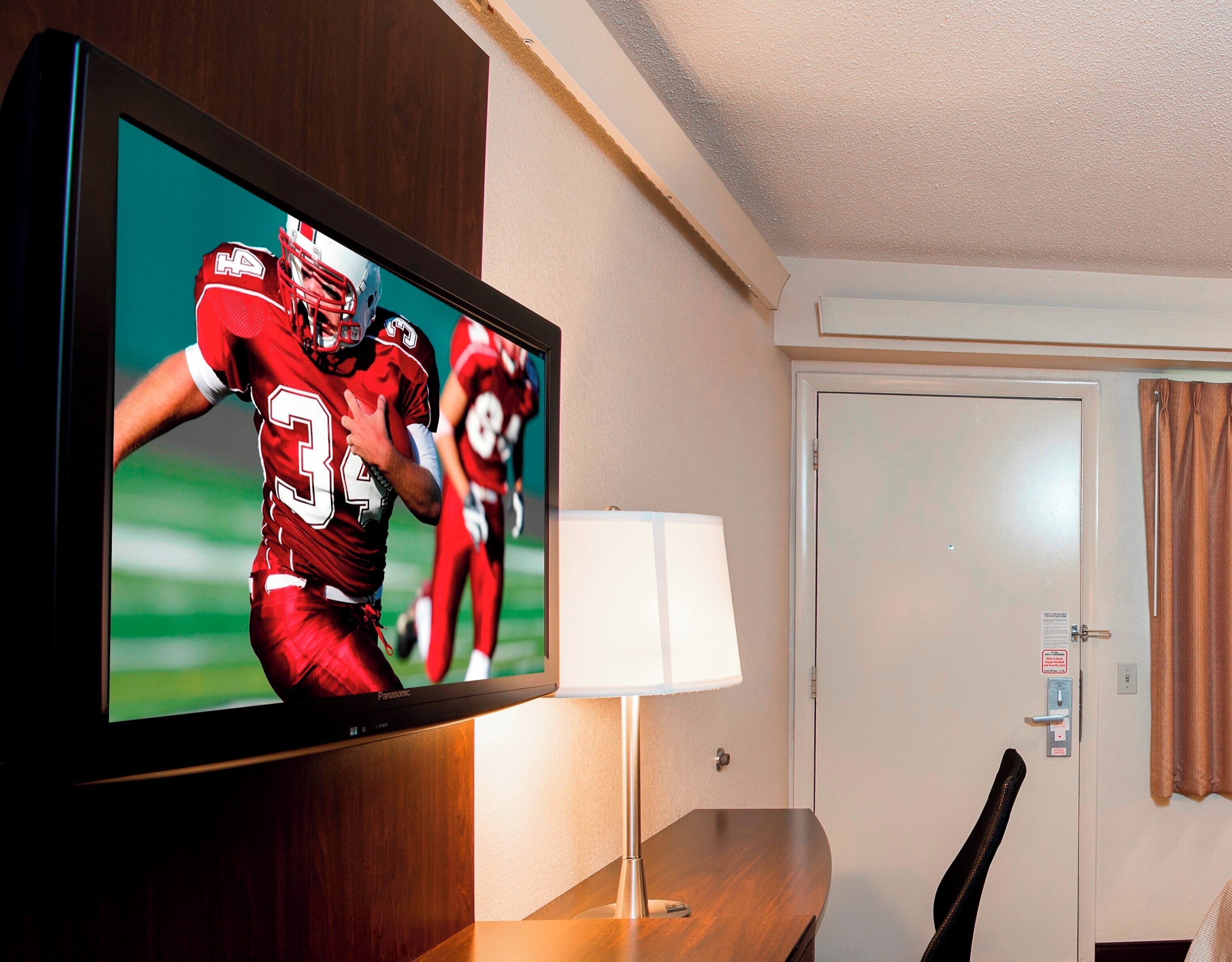 Seinälle asennettu televisio, jossa näkyy jalkapallo-ottelu, lamppu, ovi ja puinen pöytä.