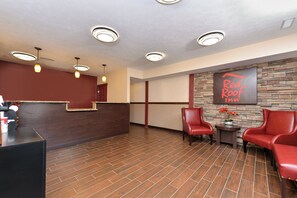 A reception area with a wooden reception desk, red chairs, a stone wall with a Red Roof Inn sign, and recessed ceiling lights. - Red Roof Inn Marietta (Marietta)