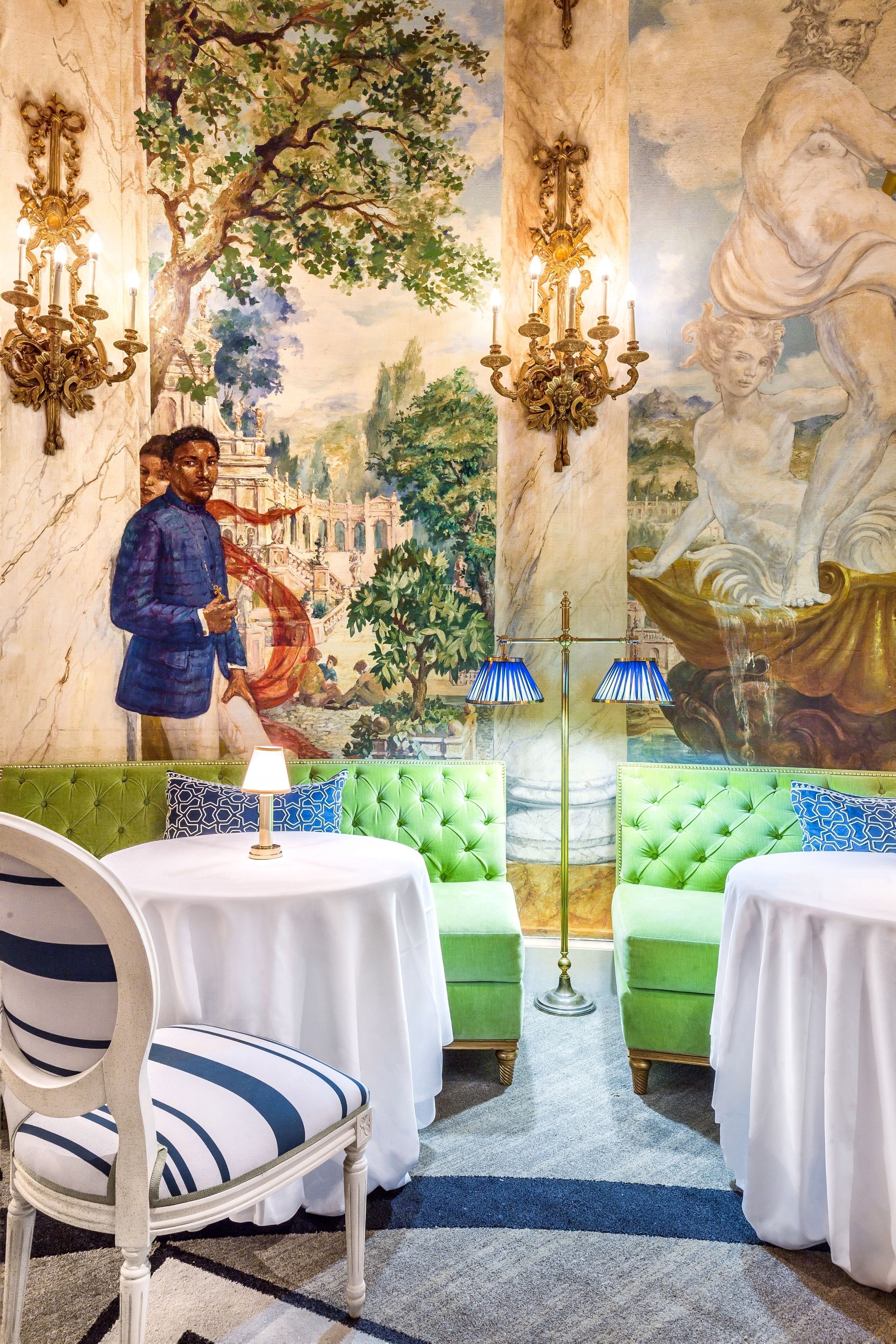 A room with a green upholstered sofa, a white table with a lamp, and a mural depicting a historical scene.