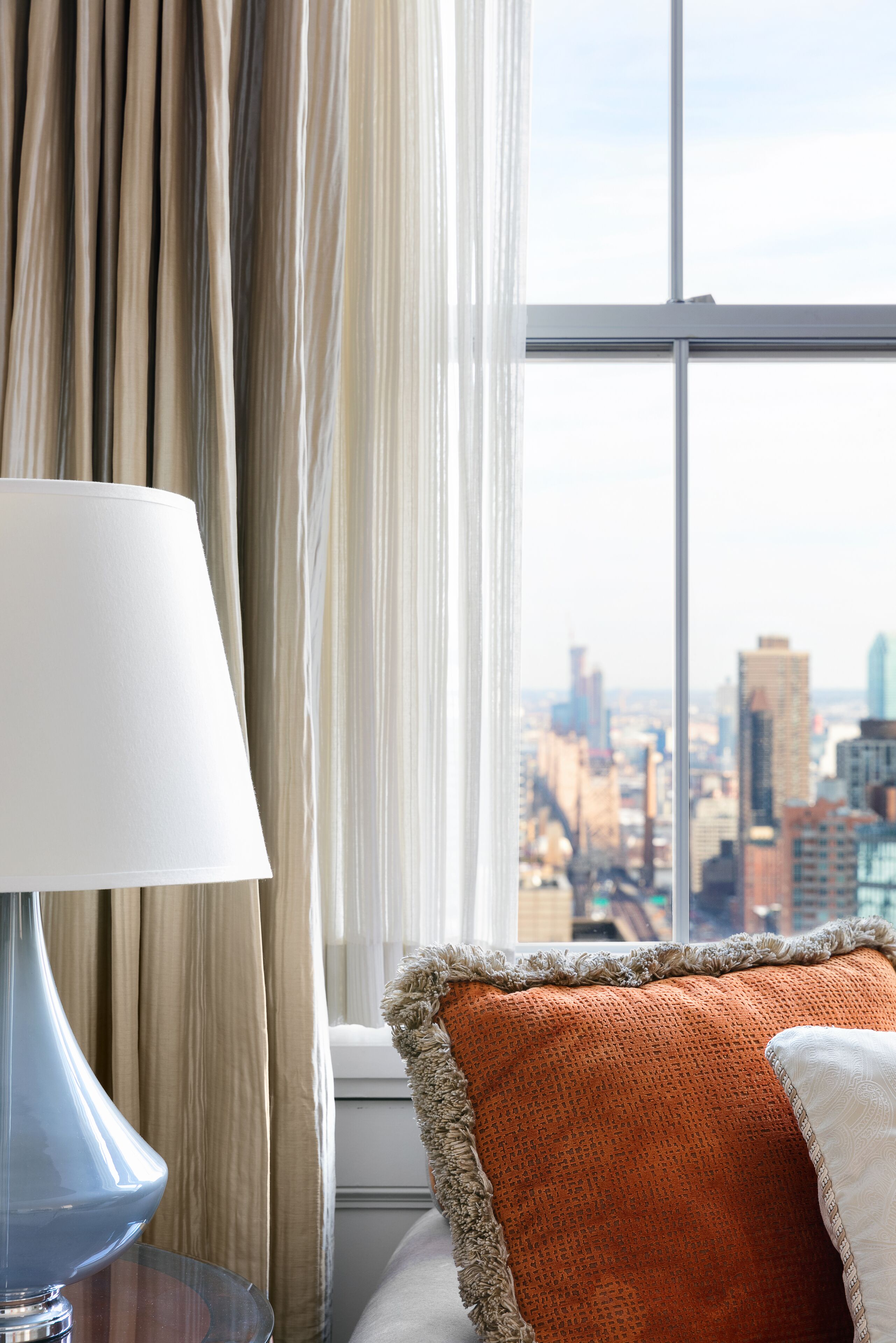 A hotel room with a large window, a lamp, and a bed with an orange pillow.