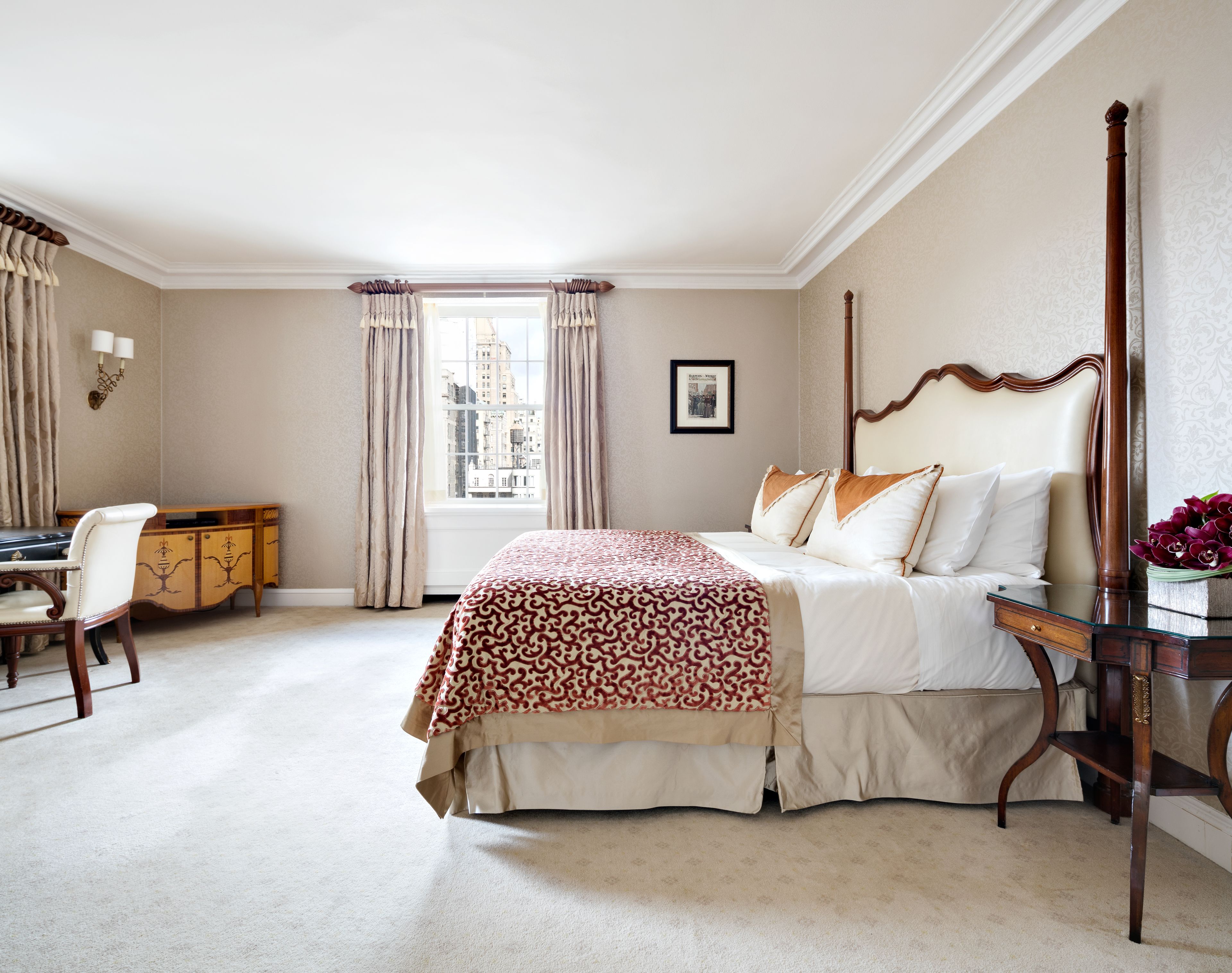 Hotelkamer met een groot bed, een bureau en een stoel. Er is een raam met gordijnen, een ingelijste foto aan de muur en een bloemenvaas op een bijzettafeltje.
