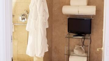 A bathroom with beige tiles, a white robe hanging on a rack, a toilet with a white seat cover, and a TV on a stand.