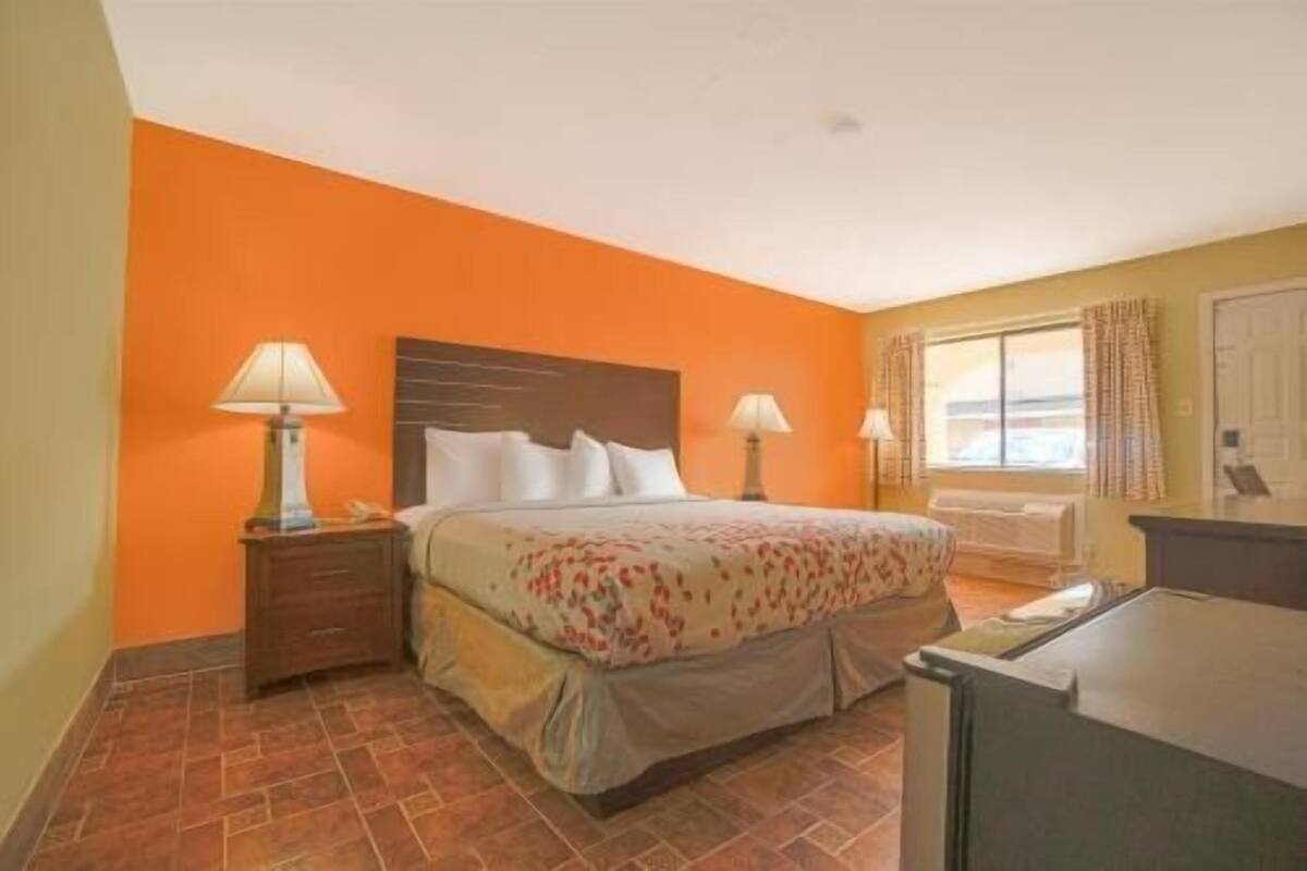 A hotel room with a bed, two bedside tables with lamps, a window with curtains, a radiator, and a small desk with a chair.