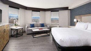 A modern hotel room with a large bed, a sofa, a coffee table, and a view of mountains through large windows. - DoubleTree by Hilton Denver - Thornton (Thornton)