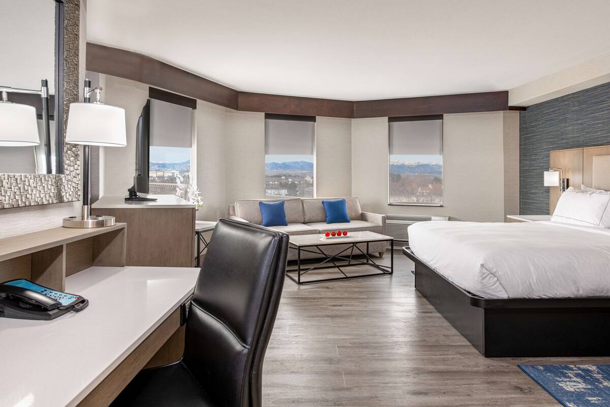 A modern hotel room with a desk, a sofa, a bed, and large windows offering a city view.