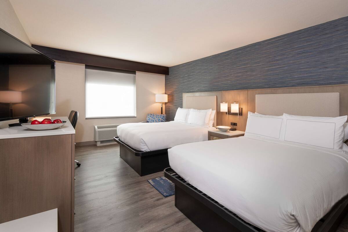 A hotel room with two beds, a desk, a TV, and a window.