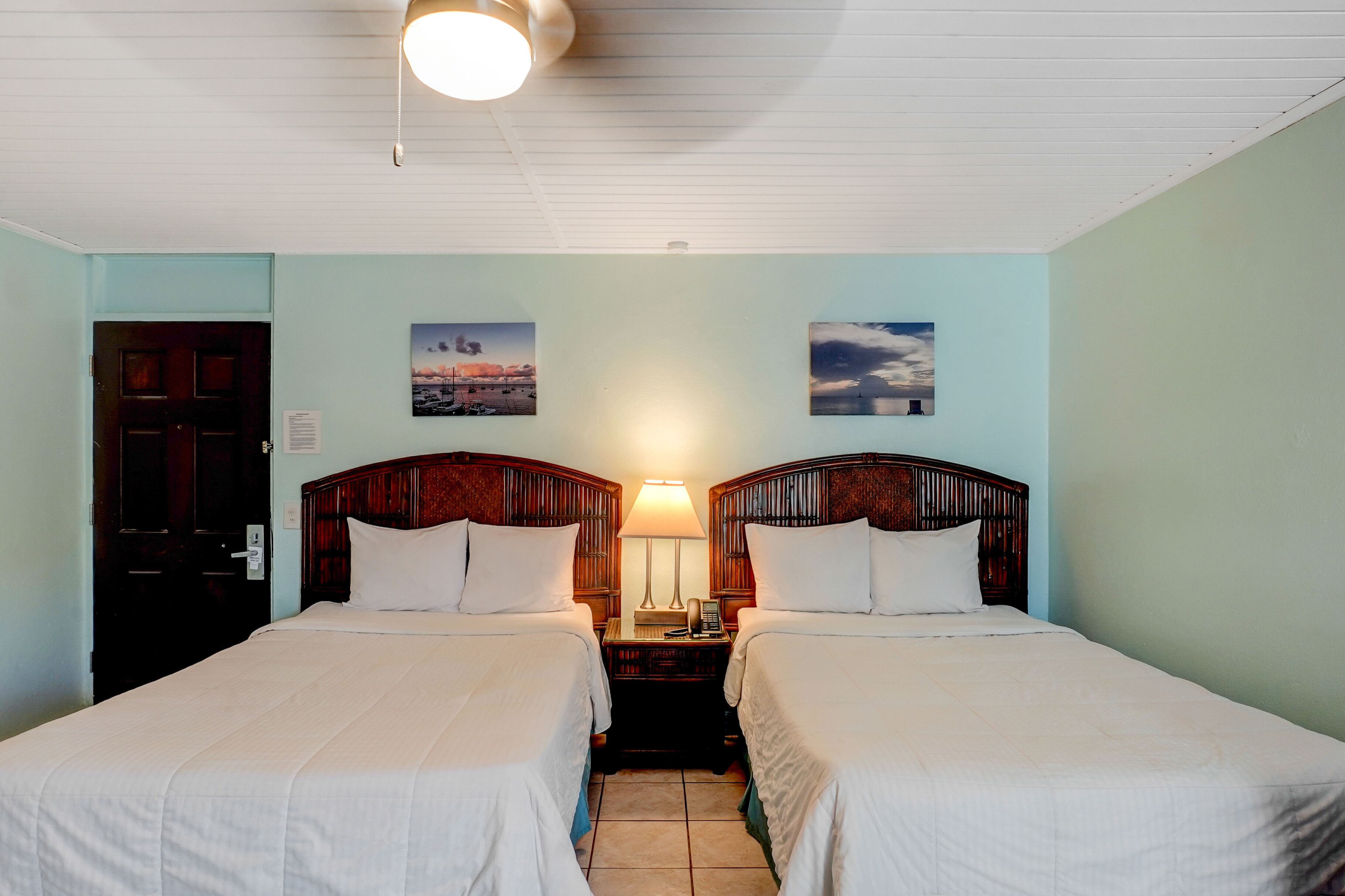 Une chambre d’hôtel avec deux lits simples, une tête de lit en bois, une table de chevet avec une lampe et deux tableaux accrochés au mur.