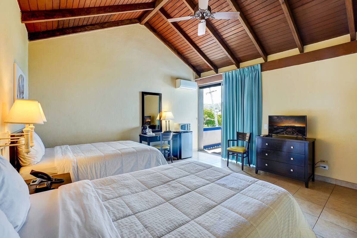 A hotel room with two beds, a desk with a chair, a television, and a ceiling fan.