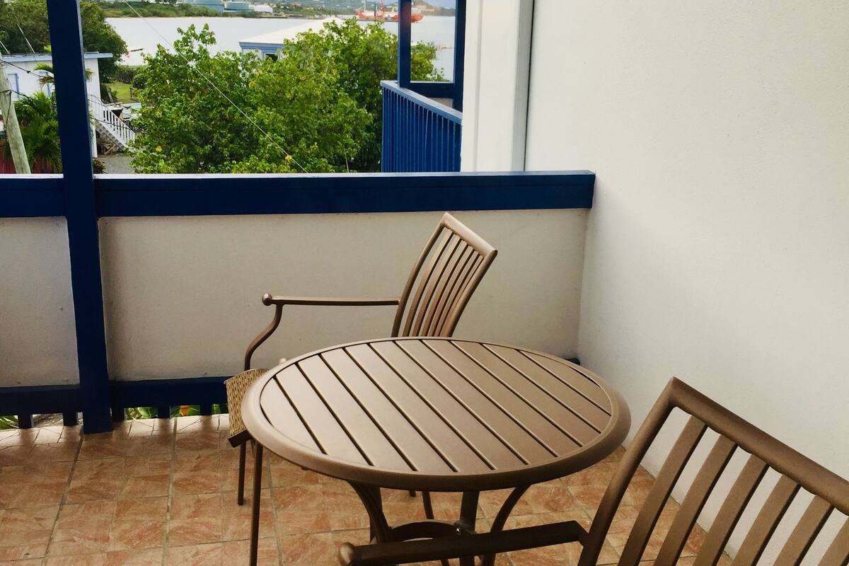 A balcony with a round table and two chairs, a view of the sea and hills, and tiled flooring.