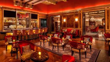 A luxurious bar area with red upholstered chairs, a bar counter with bottles, and a chandelier.