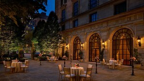Un área de comedor al aire libre con mesas y sillas, rodeada por un patio de piedra y amplias ventanas arqueadas.