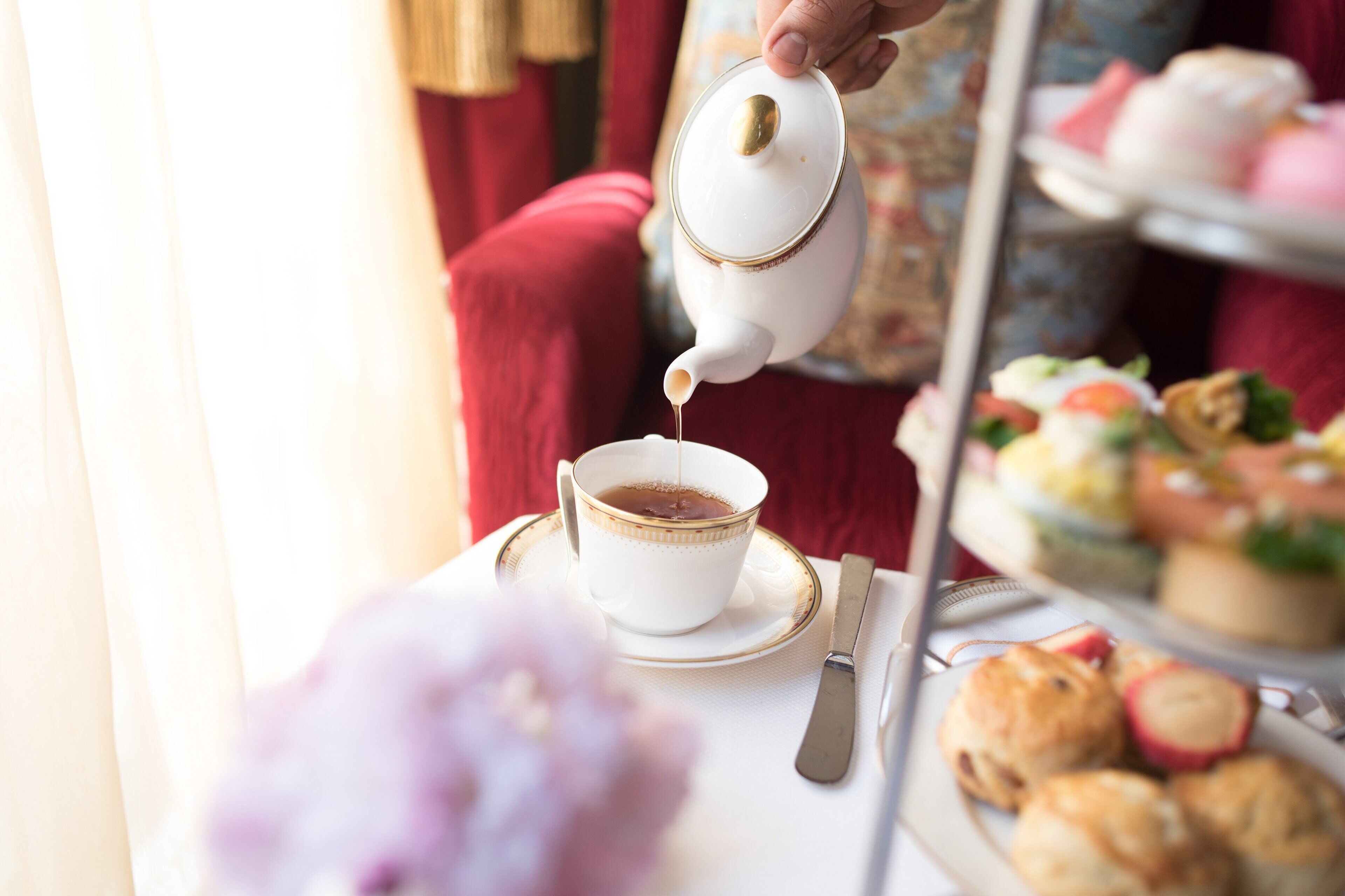 A teapot pouring tea into a cup on a table with a plate of pastries and a tiered dessert display in the background.