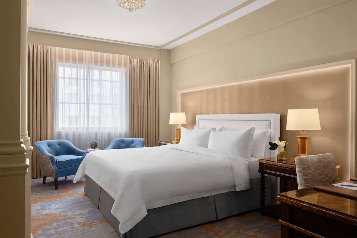 A hotel room with a large bed, two bedside lamps, a chair, a desk, and a view of the outdoors through a window with curtains.