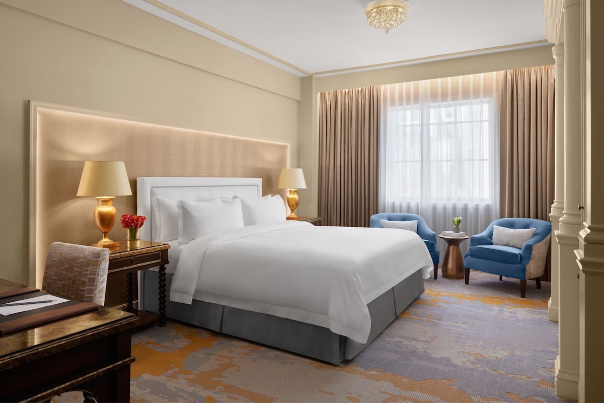 A hotel room with a large bed, two bedside lamps, a desk with a chair, a small round table with a chair, and a large window with curtains.
