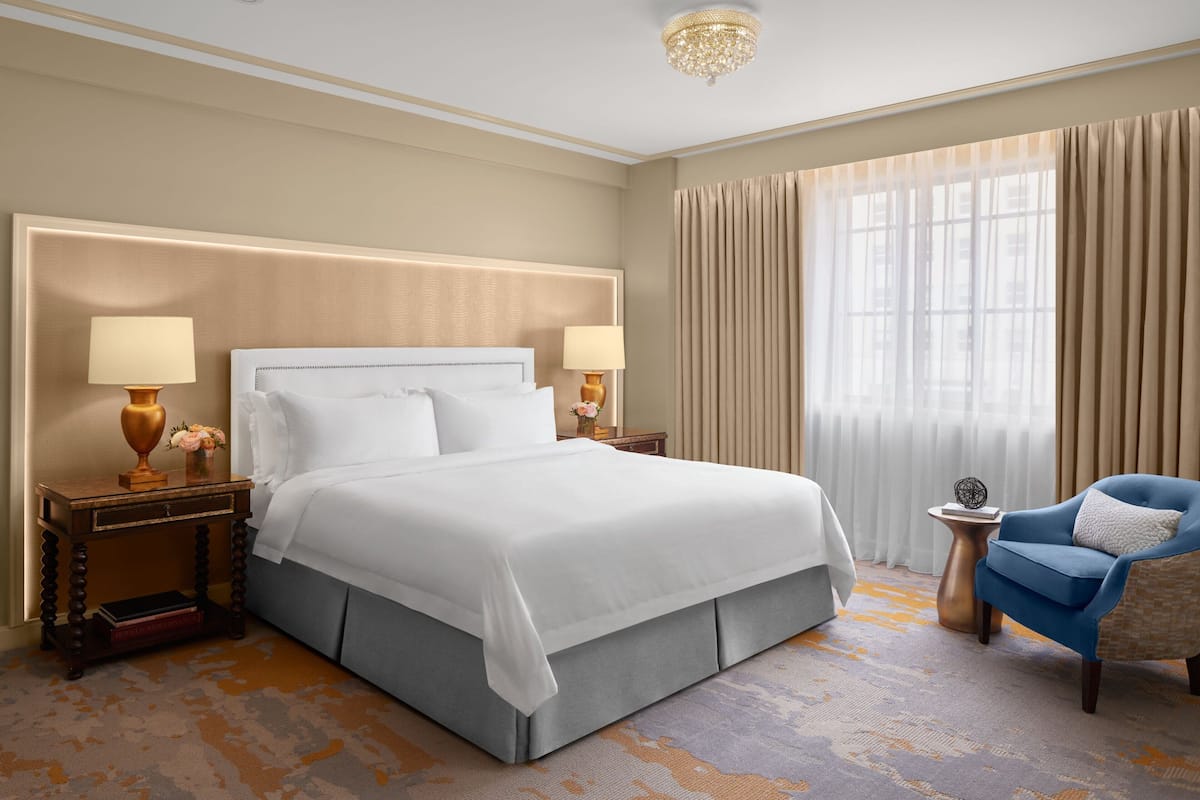 A hotel room with a large bed, two bedside tables with lamps, a chair, and a large window with curtains.