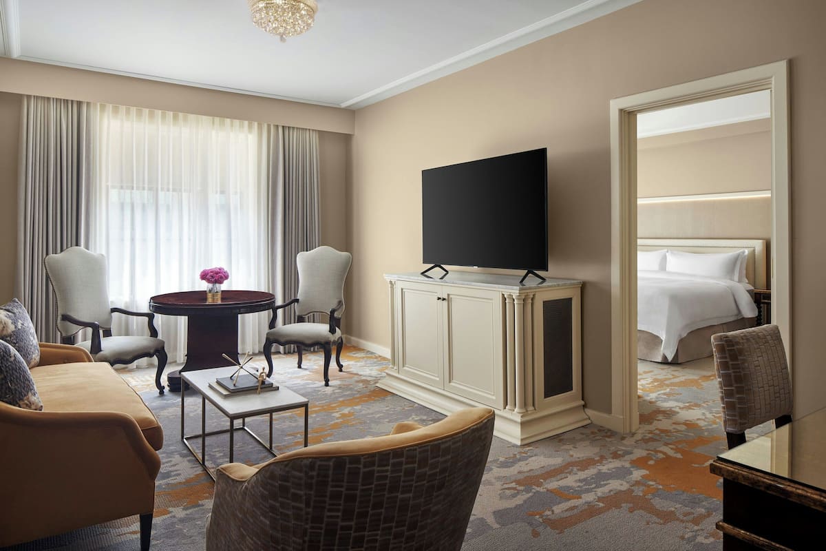 A hotel room with a sofa, two armchairs, a coffee table, a TV on a cabinet, a bed, and a large mirror.