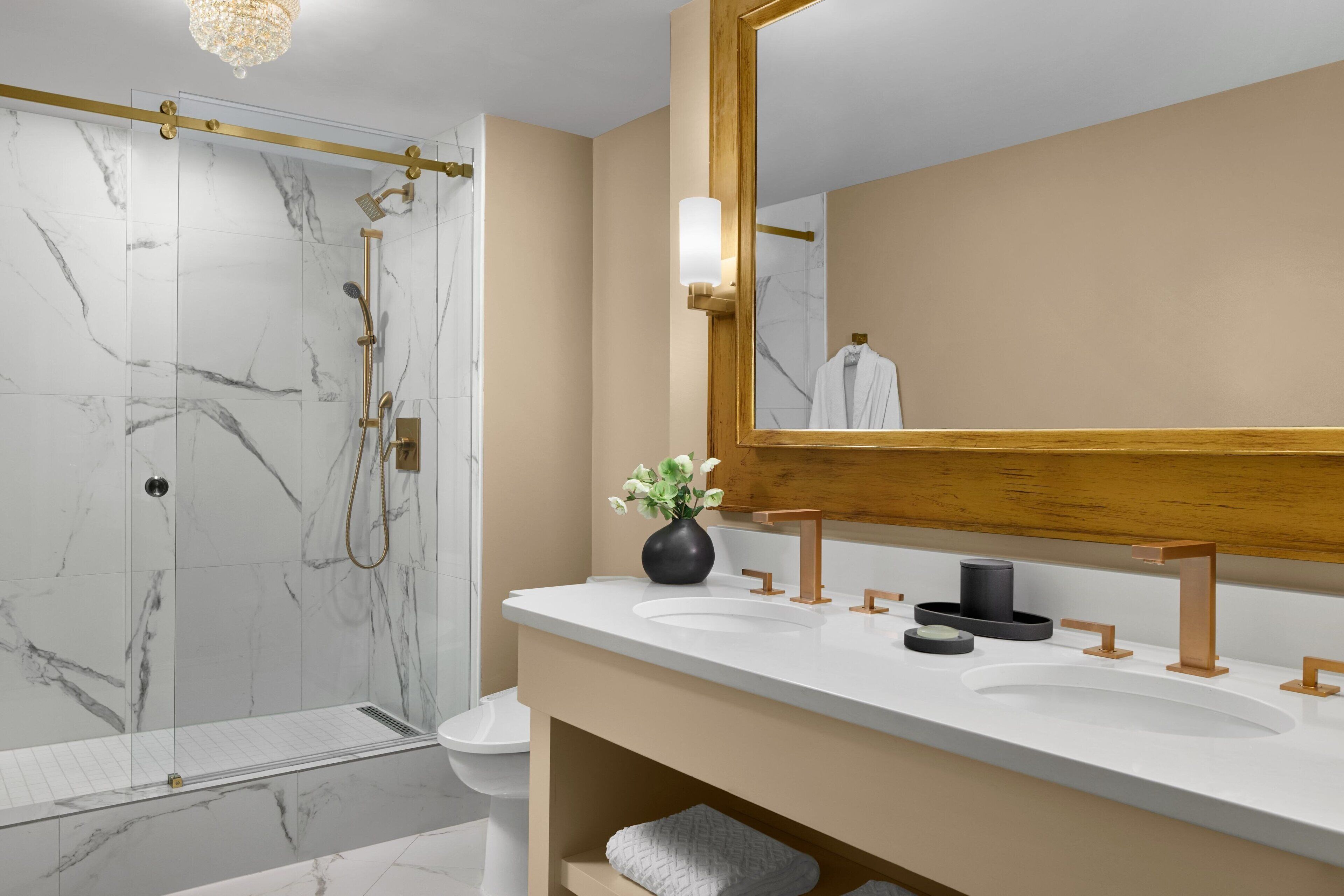 A modern bathroom with a glass-enclosed shower, a large mirror, and two sinks with gold fixtures.