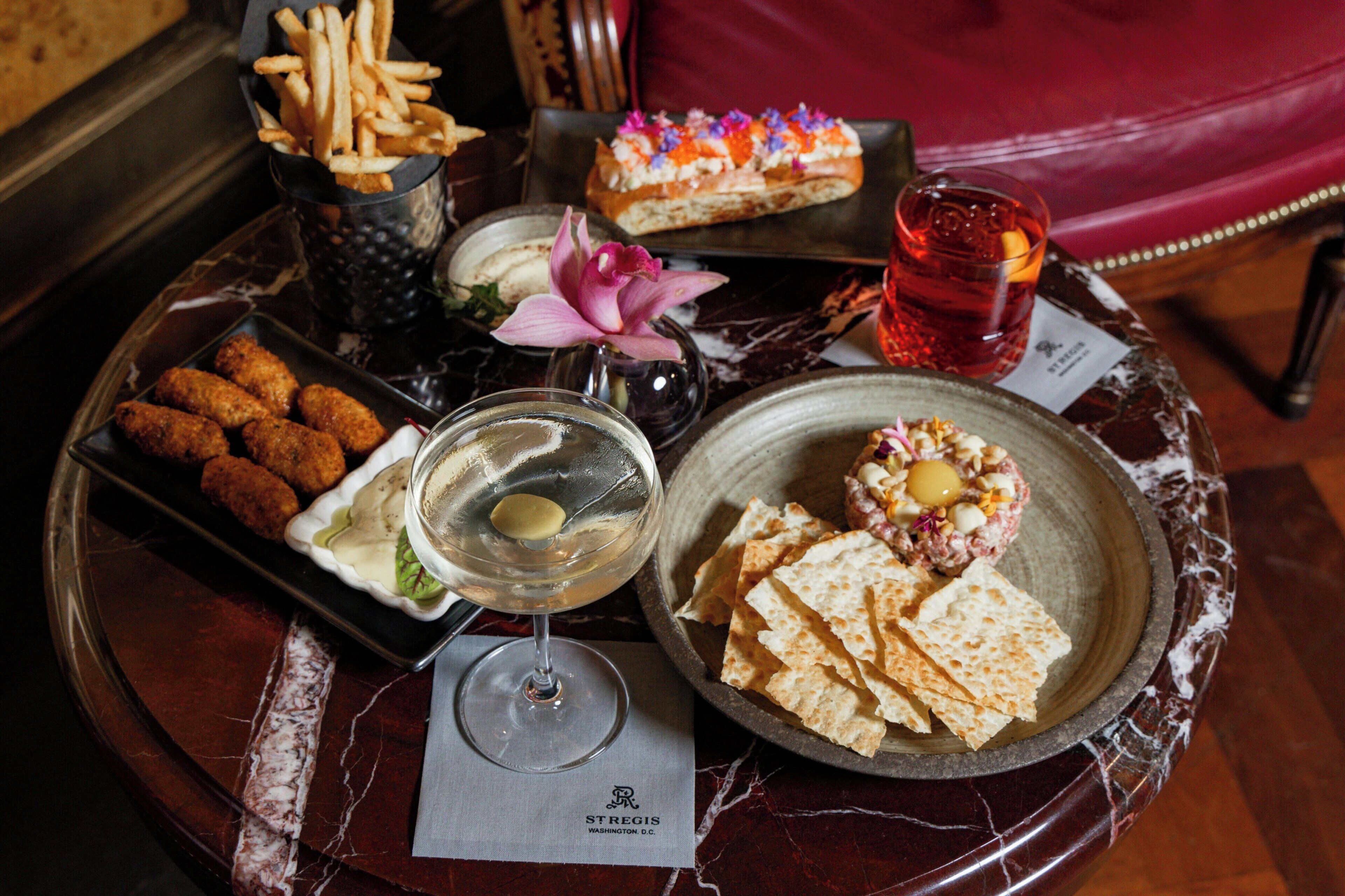 A tray with assorted food items, a glass of white wine, and a dessert on a marble table.