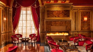 Un lobby lujoso con sillones tapizados en rojo, una chimenea de mármol y un amplio ventanal con cortinas rojas.