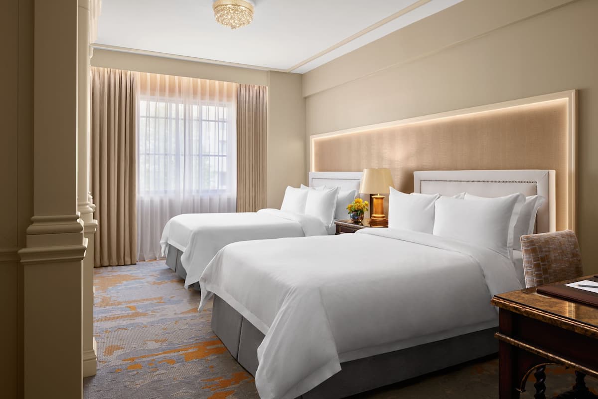 A hotel room with two beds, a large window with curtains, a chandelier, and a wooden nightstand with a lamp.