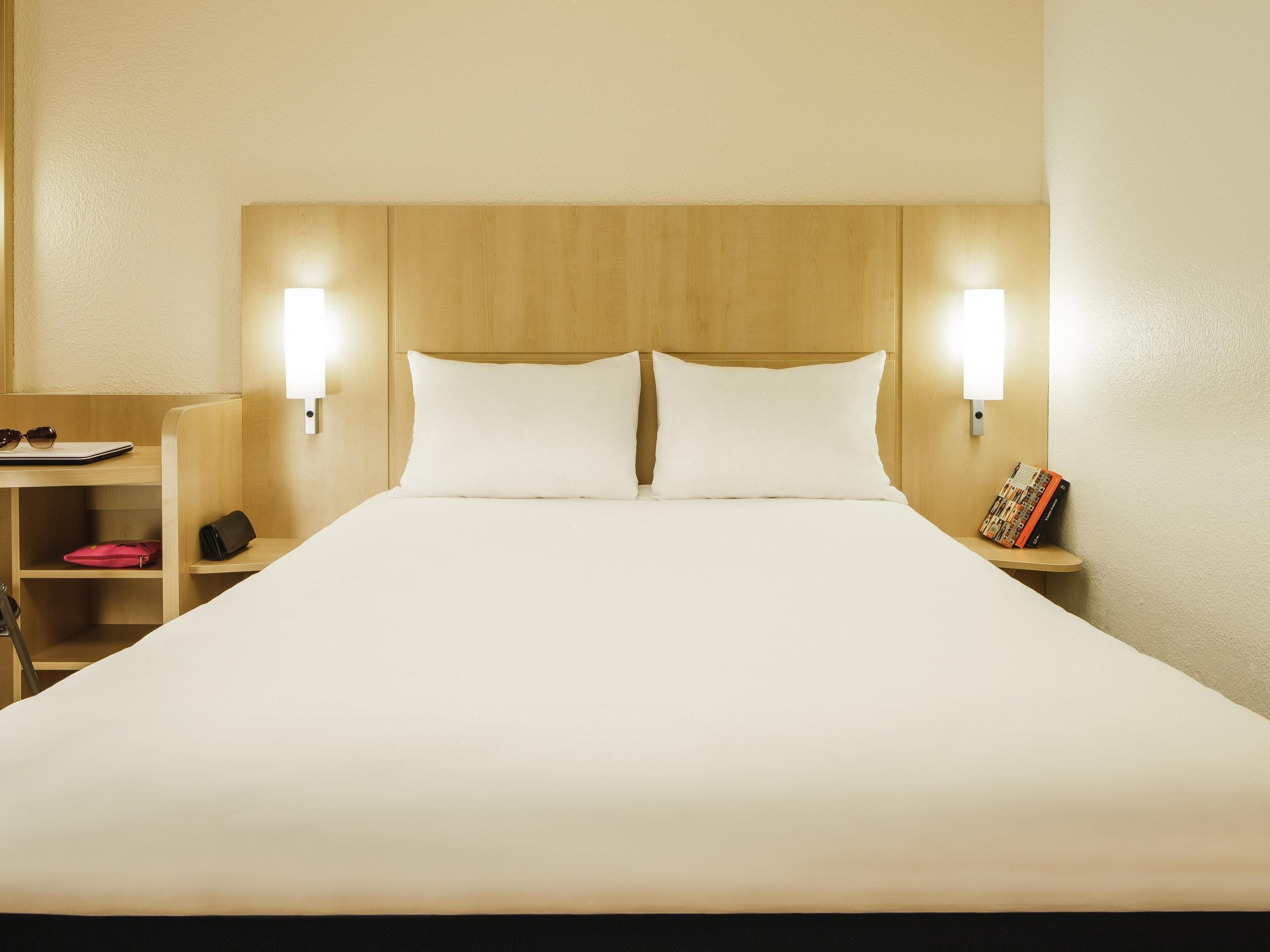 A hotel room with a large bed, two bedside lamps, a nightstand with a book, and a desk with a laptop and sunglasses.