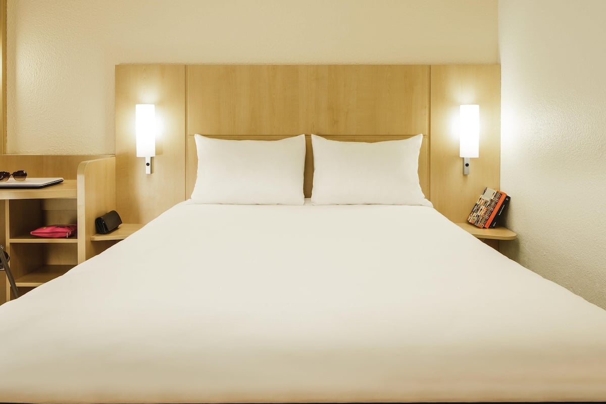 A hotel room with a large bed, two bedside lamps, a nightstand with a book, and a desk with a laptop and sunglasses.