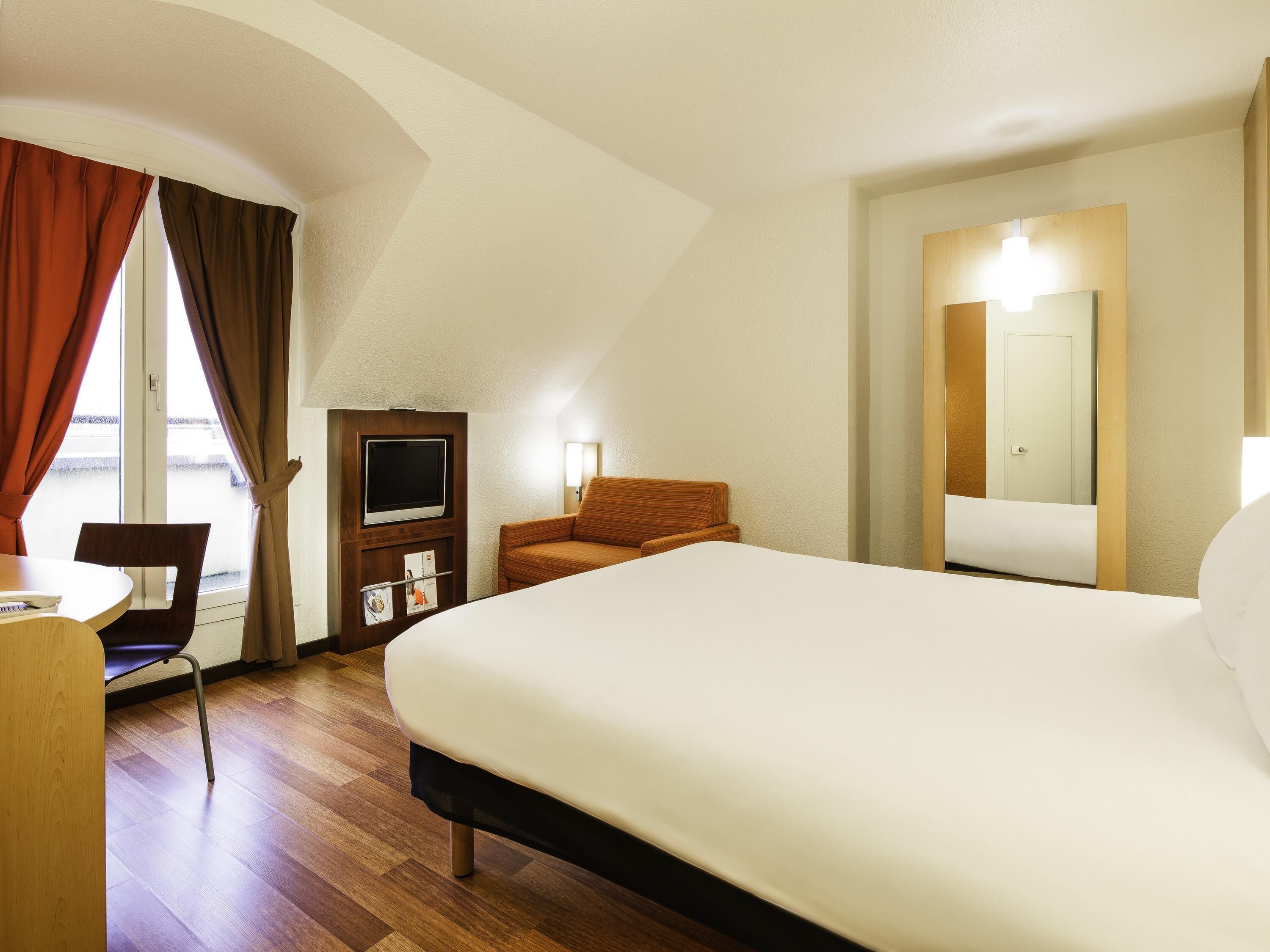 A hotel room with a large bed, a desk with a chair, a television, and a mirror.