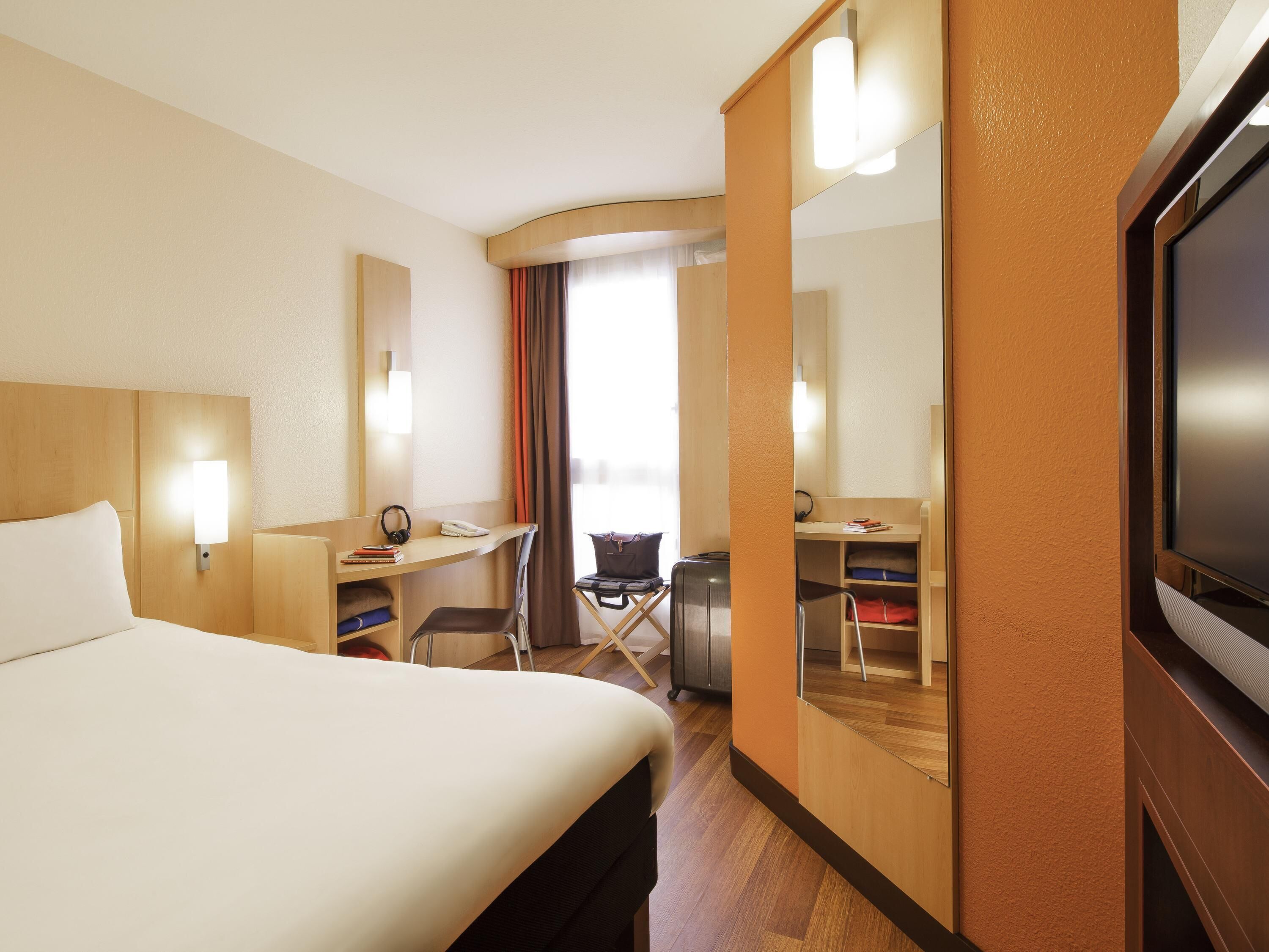 A hotel room with a bed, desk, chair, TV, and a large mirror.